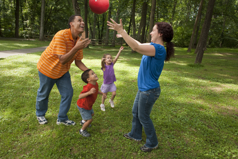 Two adults and two kids play with a ball in a grassy area with trees behind them